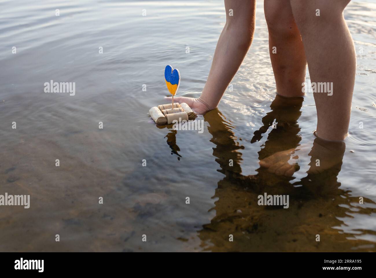 child, standing in a river, launches a home-made boat made of wine corks, instead of sail, a yellow-blue heart in colors of Ukrainian flag. Symbol of Stock Photo