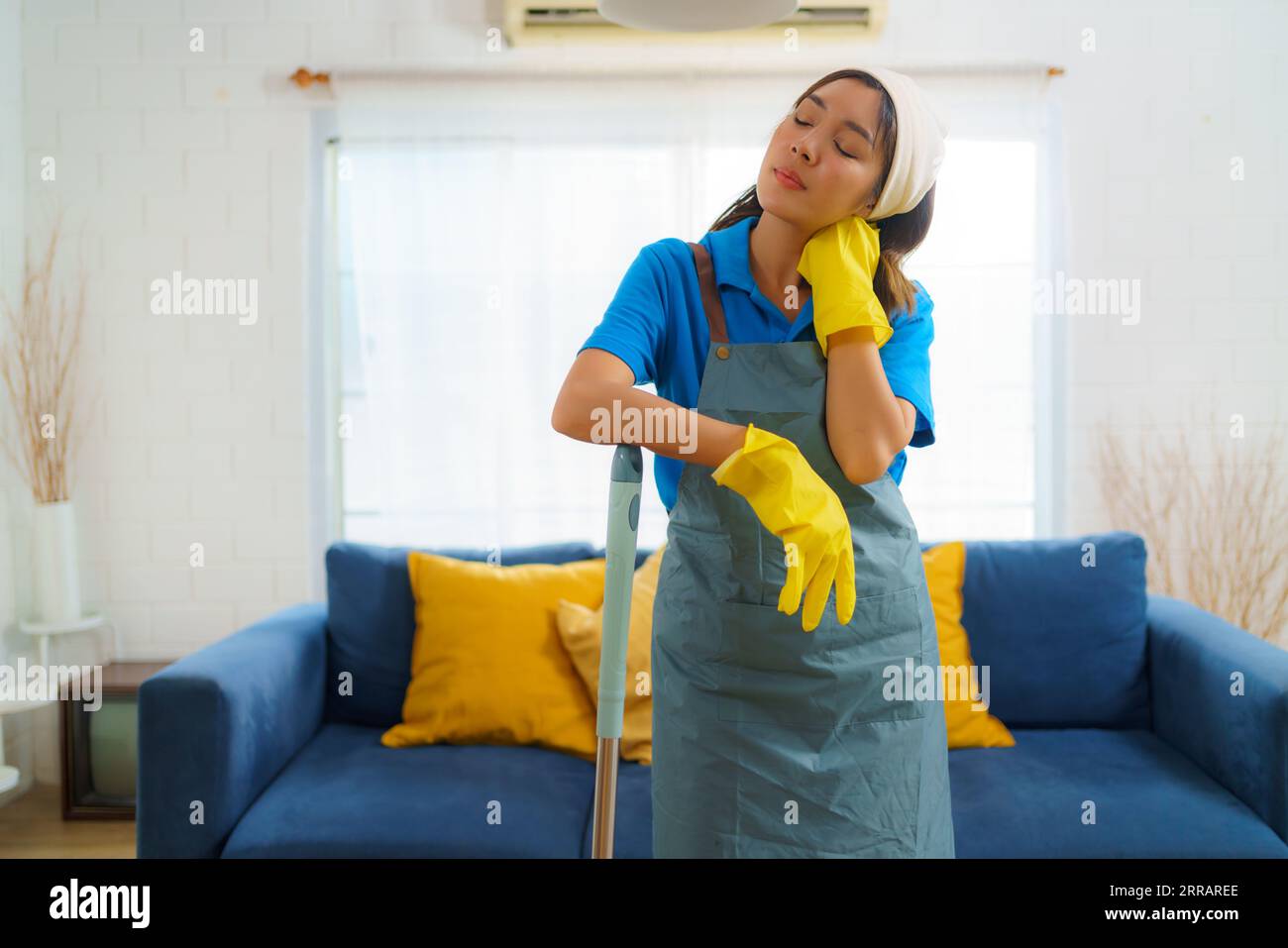Determined Asian maid battles weariness, mop in hand. Every stroke tells a story of resilience and commitment in the pursuit of cleanliness. Stock Photo