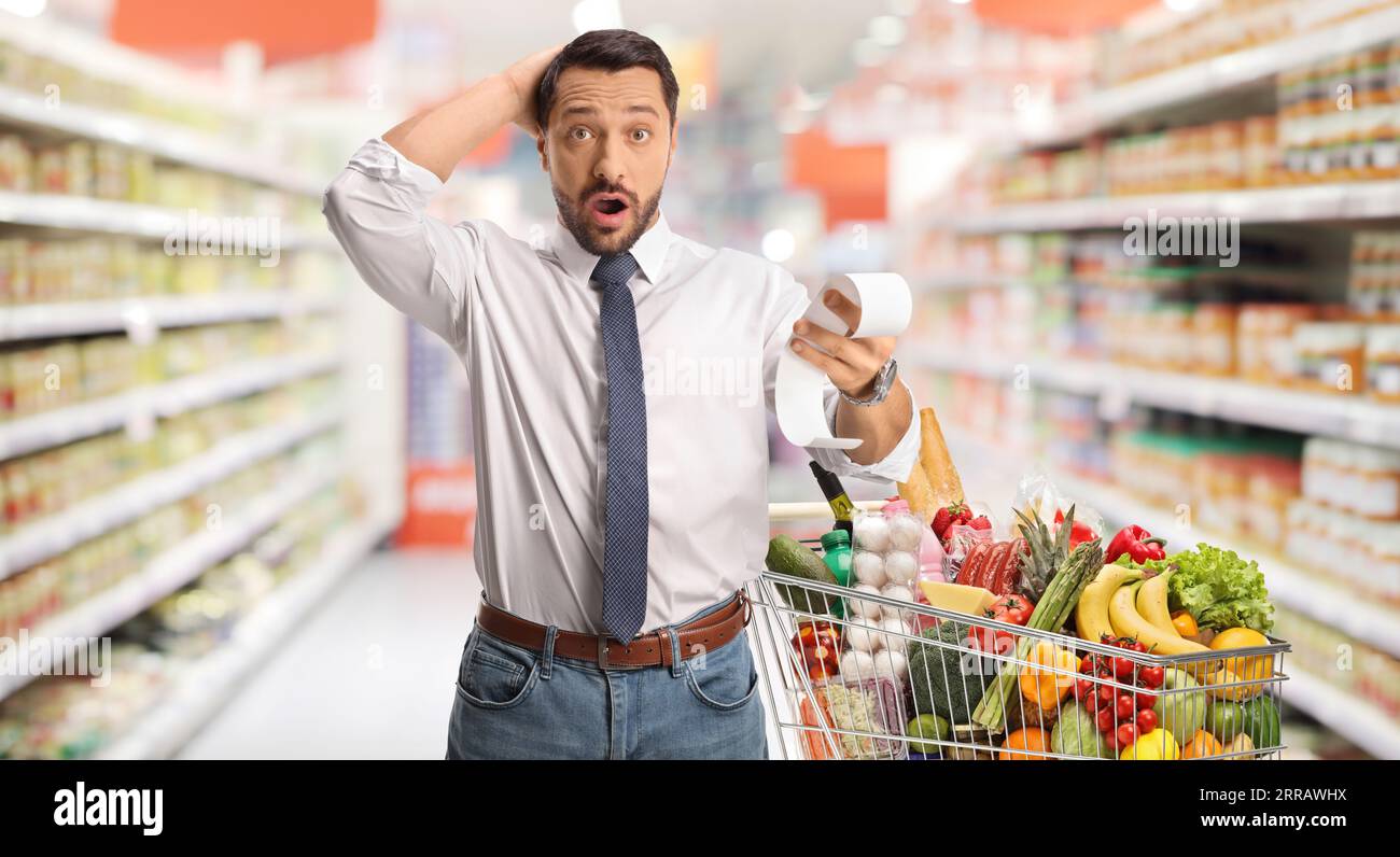 Shocked man with a bill and shopping cart holding his head in disbelief at a supermarket Stock Photo