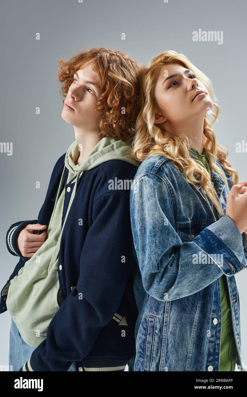 dreamy teen boy and girl in stylish casual wear standing back to back and looking away on grey Stock Photo