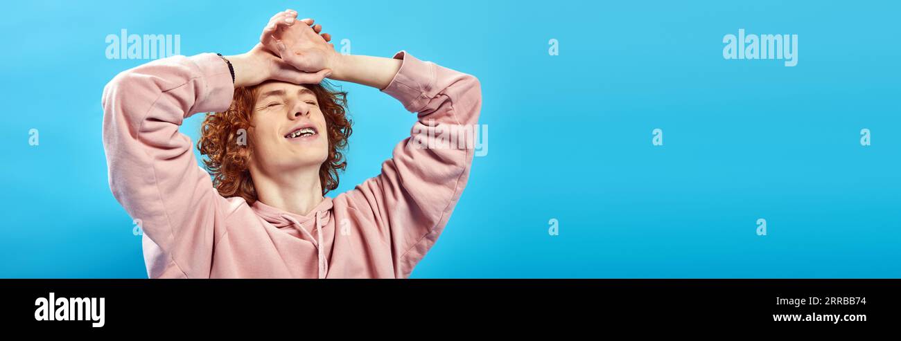 dreamy, smiling redhead teen boy in hoodie with closed eyes and hands on forehead on blue, banner Stock Photo