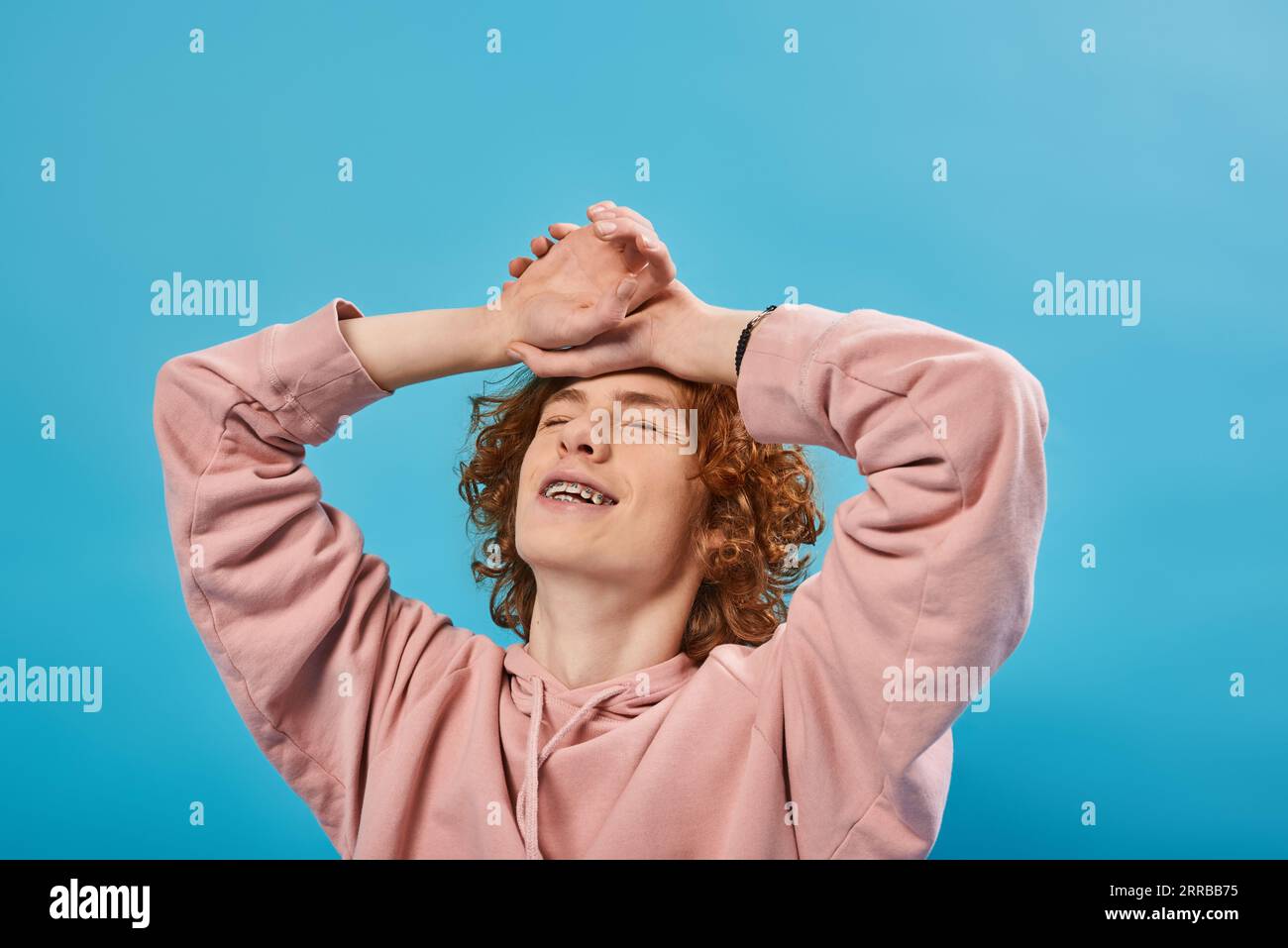 positive, dreamy redhead teen guy in hoodie standing with closed eyes and hands on forehead on blue Stock Photo
