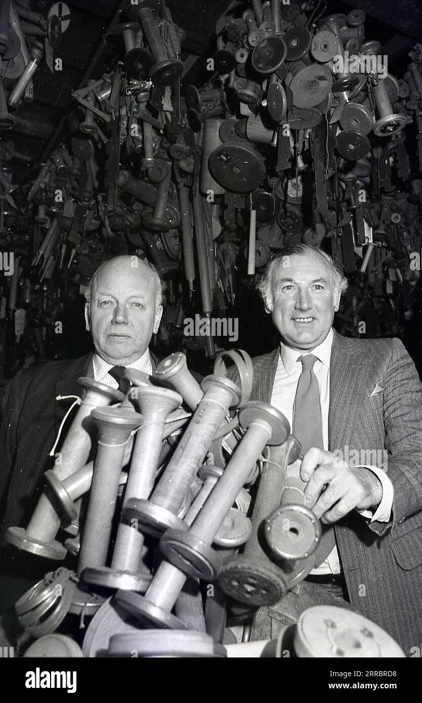 1983, two suited mature men holding large spindles or bobbins used to hold yarn or thread, sitting inside a building, Yorkshire, England, UK, the last makers of bobbins in Britian. Behind them handing up a large selection on different spindles. Stock Photo
