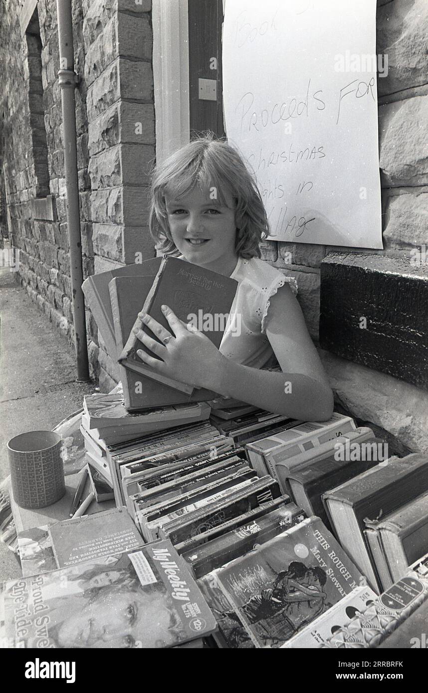 1983, historical, a young girl selling second-hand books from a stall outside a viilage stationery shop, Yorkshire, England, UK to try and raise money for christmas lights in her village. Stock Photo