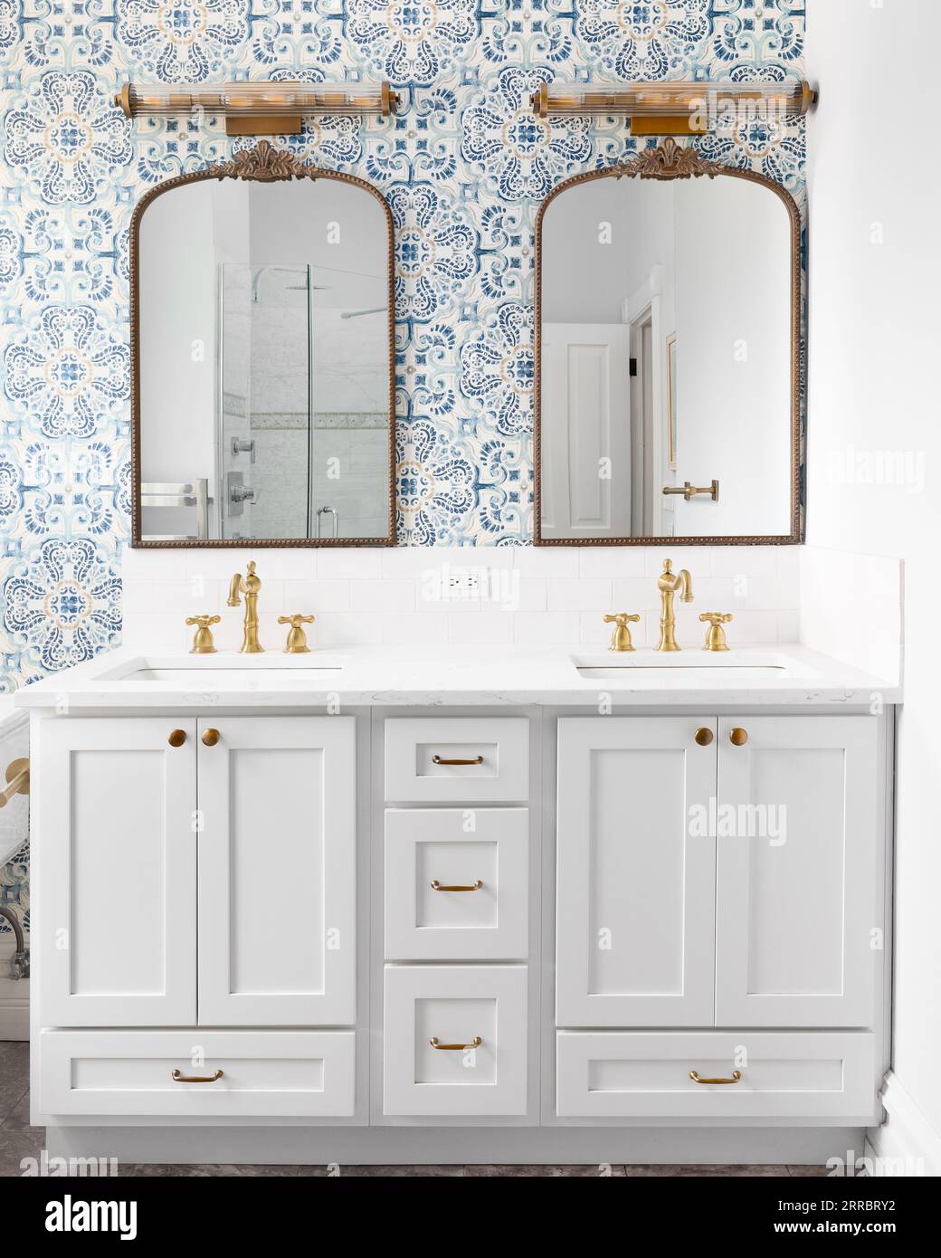 A bathroom detail with a blue and yellow pattern wallpaper, bronze mirrors and light fixtures, gold faucets, grey cabinet and subway tile backsplash. Stock Photo