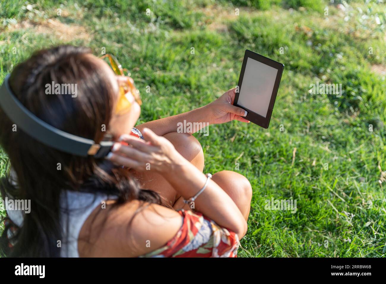 Woman with headphones holding a e-book with blank screen while sitting on grass in a park. Technology concept. Stock Photo