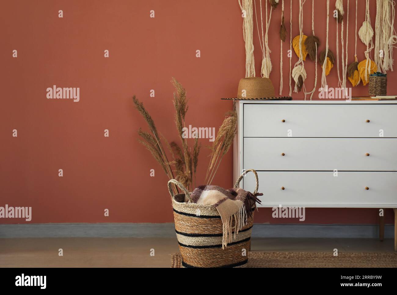 Straw hat on chest of drawers, basket and pampas grass near color wall