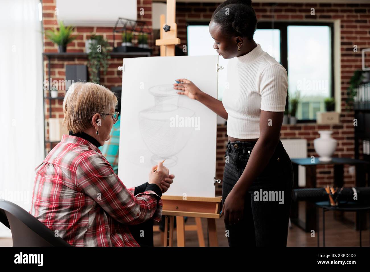 Senior woman engaging in social activities, taking drawing lesson. African American young woman art instructor teaching older adults to draw in community center standing near easel assisting student Stock Photo