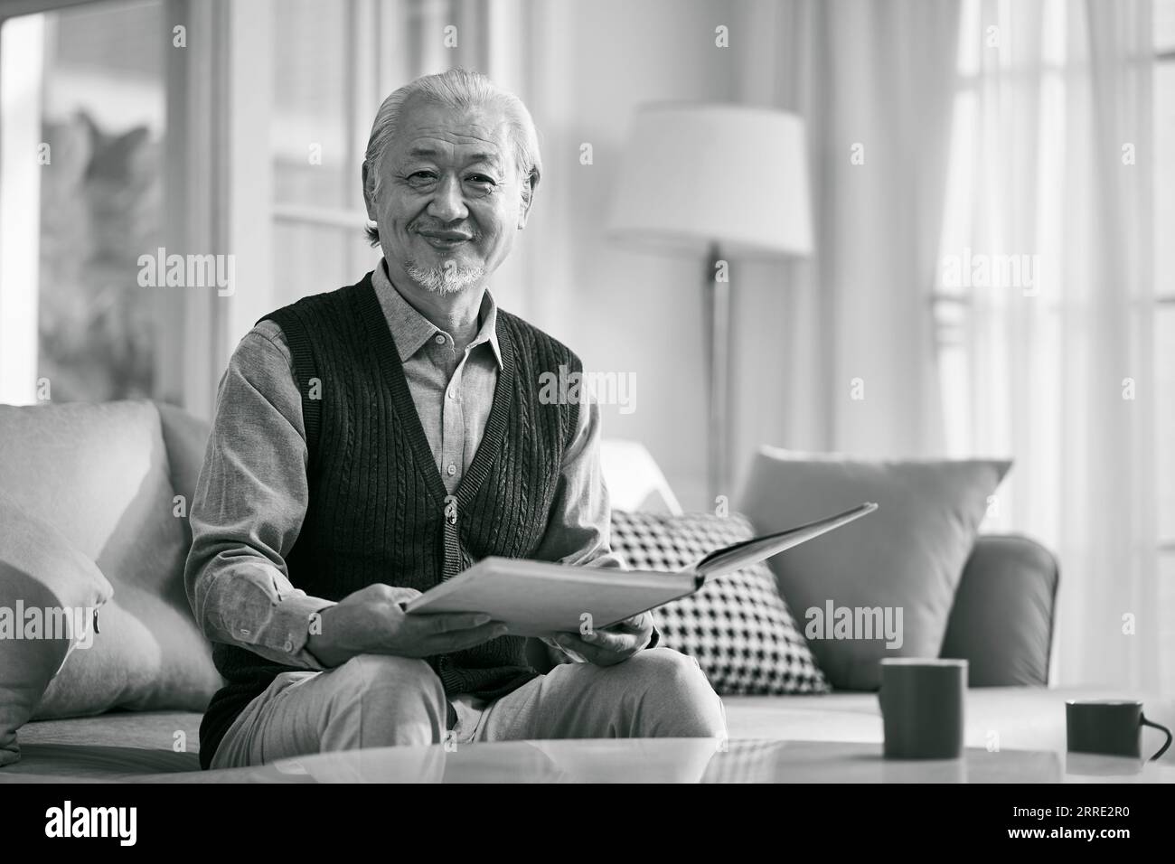 happy asian old man sitting on couch at home reading a book looking at camera smiling, black and white Stock Photo