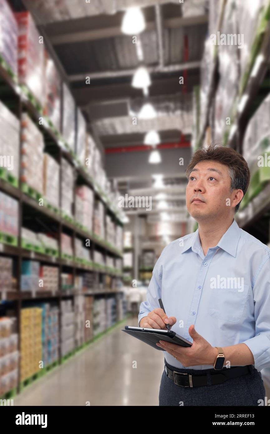 a middle-aged Asian businessman holding a tablet at a warehouse full of goods Stock Photo
