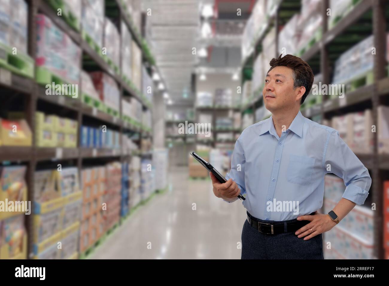 a middle-aged Asian businessman holding a tablet at a warehouse full of goods Stock Photo