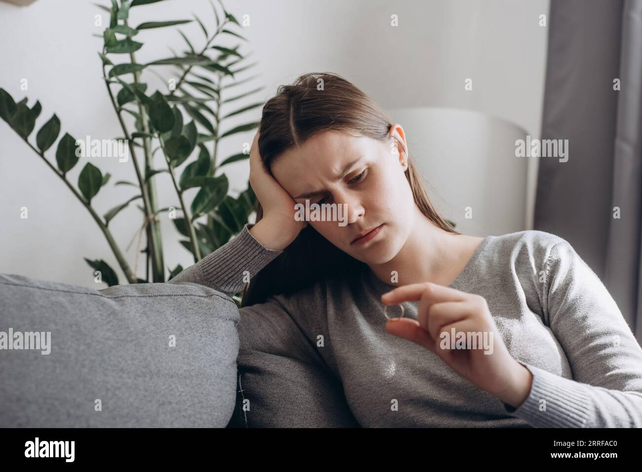 Frustrated unhappy desperate woman sits on sofa holding engagement ring in hand, stressed young female suffering from relations breakup or fiancee bet Stock Photo