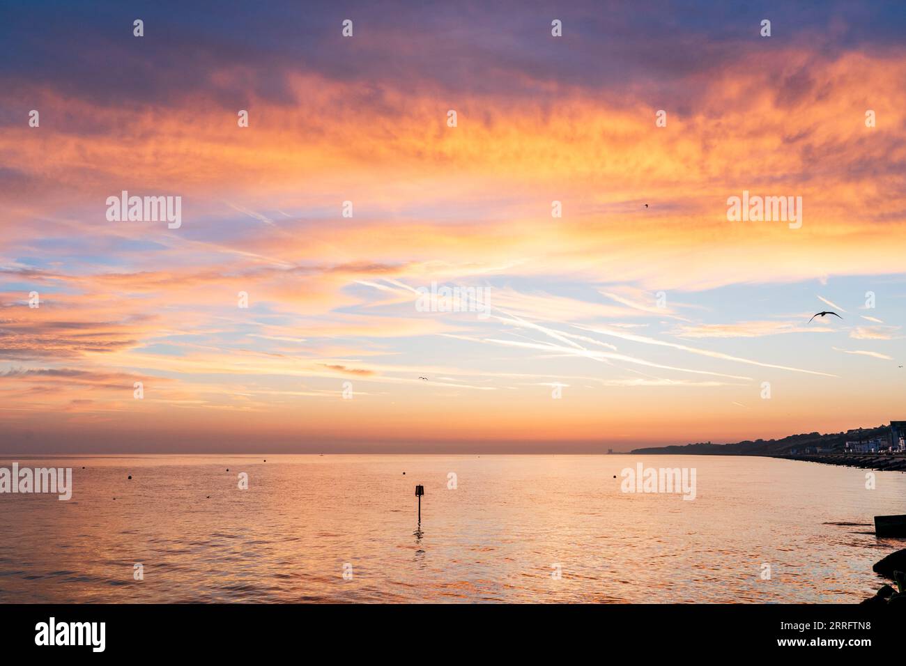 The dawn sky over the sea at Herne Bay on the North Kent coast. Orange and yellow clouds against a blue sky with a band of orange on the horizon. The clam seea reflecting he orange colour. Stock Photo
