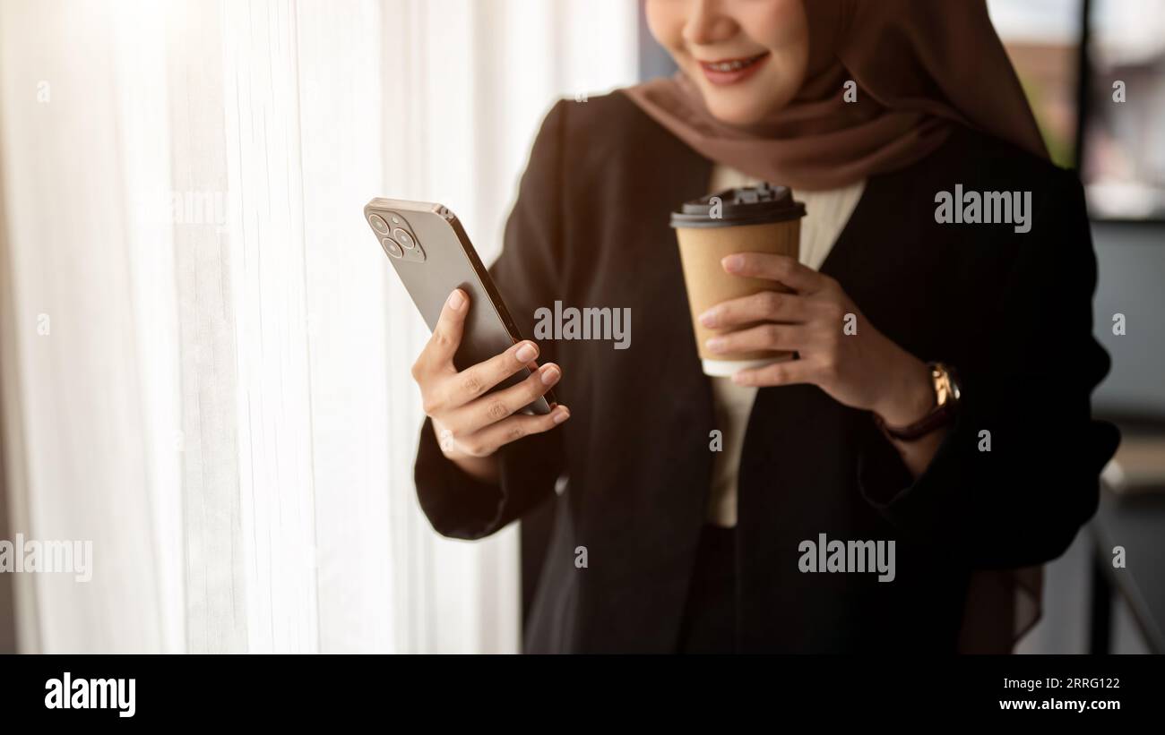 Cropped image of a happy and smiling Asian Muslim businesswoman in a formal black suit and hijab is checking messages on her smartphone and having cof Stock Photo
