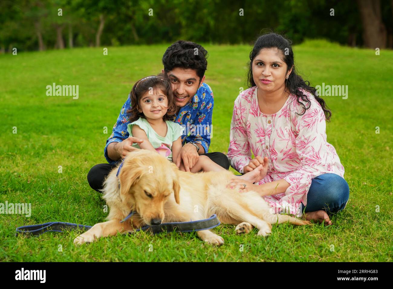 Happy young indian family having fun together at summer park. Mother, father and daughter with labrador dog in garden. Stock Photo