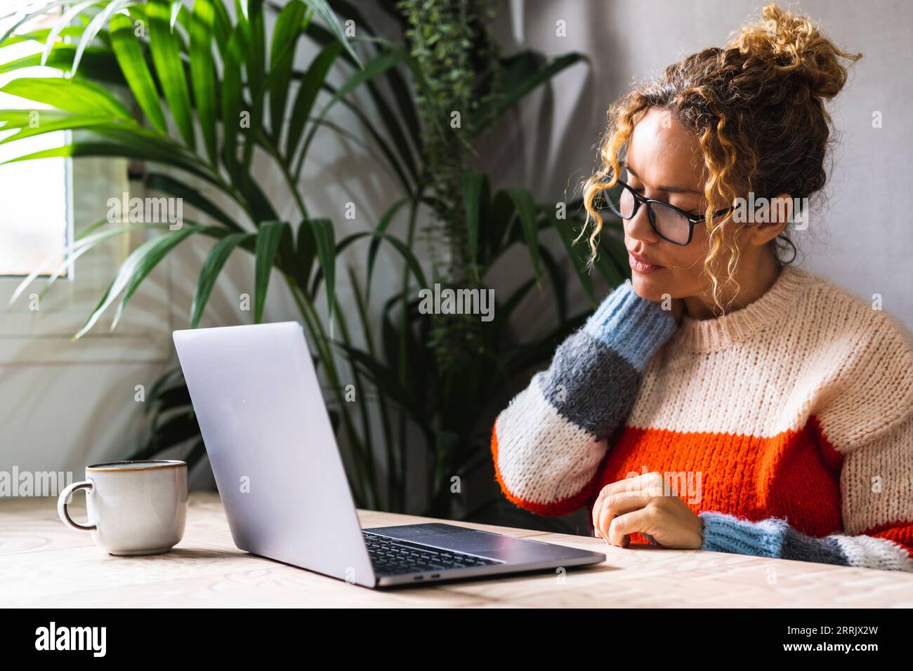One adult woman working on laptop and touching painful her neck and shoulder for bad posture sitting at the table workplace. Female people massage body while use computer. Tired and stressed lady Stock Photo