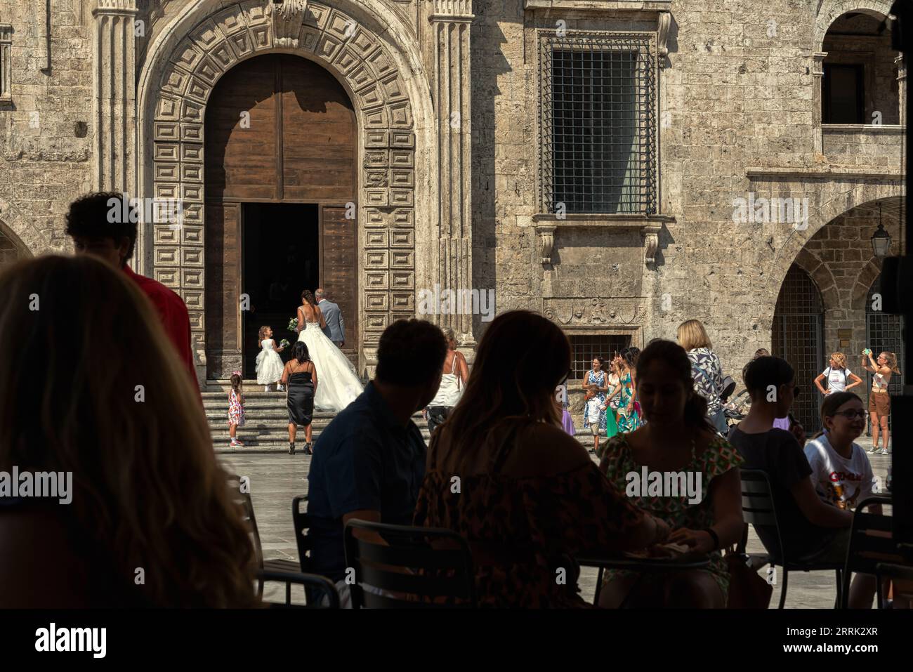 Tourists sitting at the café under the arcades in Piazza del Popolo admire the bride entering the Palazzo dei Capitani for the wedding. Ascoli Piceno, Stock Photo