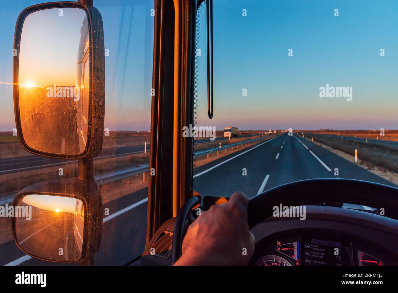 View from the driver's seat of a truck, with a straight road ahead and the sun rising. Stock Photo