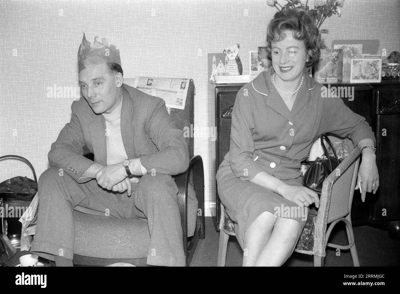 England. c.1960 – A man and a woman are sitting in a lounge watching television on Christmas Day. The man is wearing a tweed suit with a sweater and a tissue paper hat from a Christmas cracker. He is sitting in an armchair with a newspaper draped over the back. The woman is wearing a two-piece suit and a string of pearls and is sitting in a Lloyd Loom type chair. Behind them is a dark wood sideboard cabinet with greetings cards displayed on top. To the side is a full brass coal scuttle and fire irons. Stock Photo