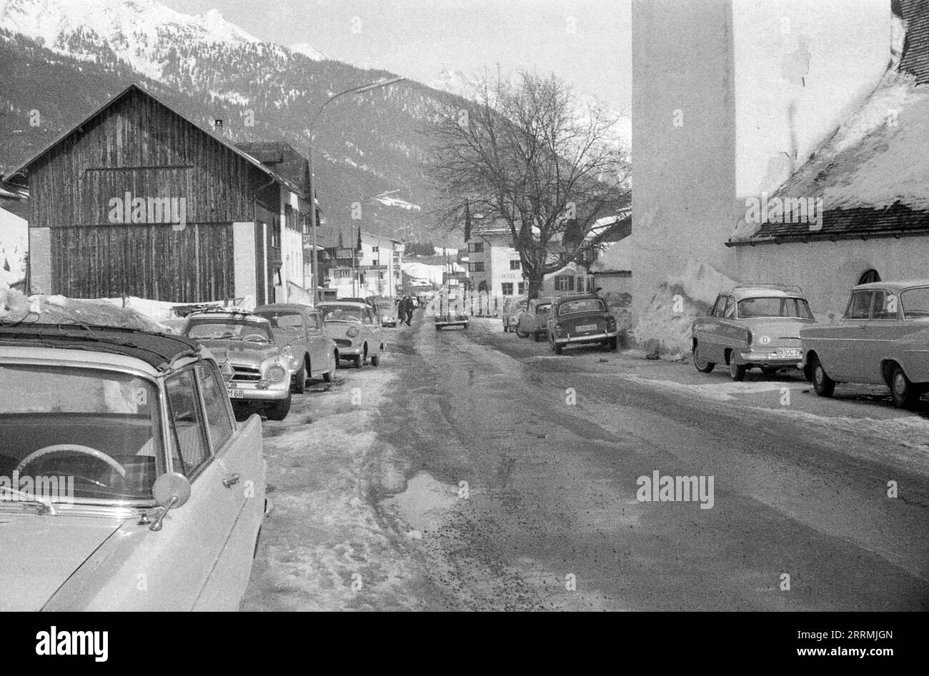 Tyrol, Austria. c.1960 – A view of Dorfstraße in the village of St Anton am Arlberg. On the right is St. Anthony of Padua church. Cars are parked along both sides of the road, which is in a slushy condition. Hotels and the snow-clad mountains of the Eastern Alps are visible in the distance. Stock Photo