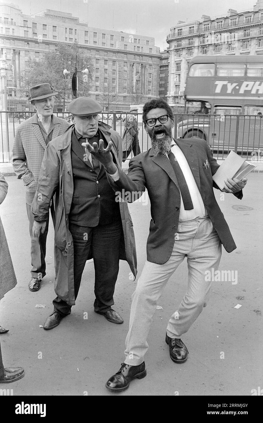 London. 1965 – An evangelist preaching at Speaker’s Corner, Hyde Park at the junction of Cumberland Gate, near Marble Arch, London. The preacher is holding a bible in one hand and is gesticulating with the other. A group of spectators have gathered around him. A double-decker London bus is passing by bearing an advertisement promoting Ty-Phoo Tea. The Cumberland Hotel and the building at 140 Park Lane are visible in the distance. Stock Photo