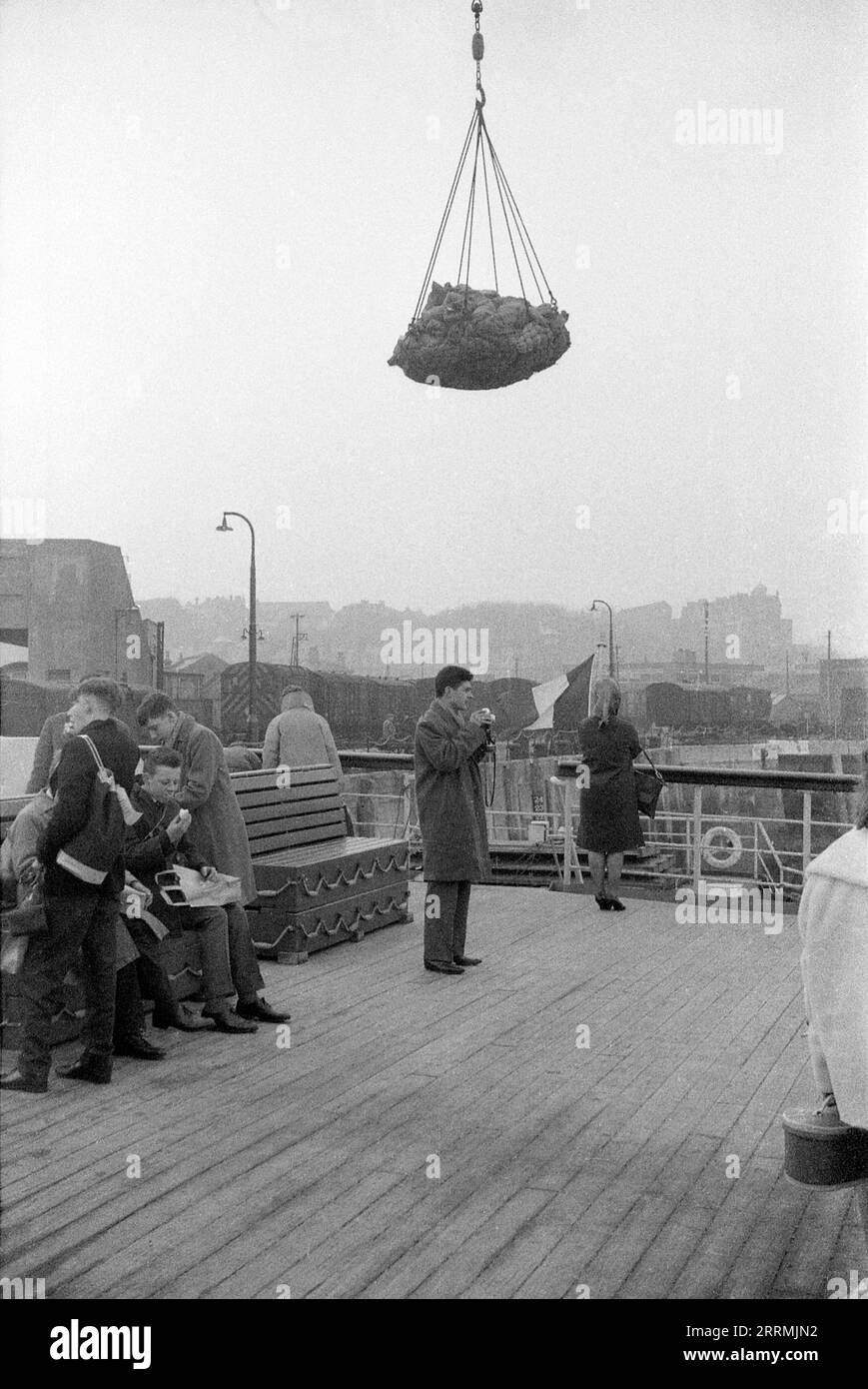 Kent. c.1960 – Passengers on the deck of a French cross-channel ferry at Folkestone Harbour, next to the Folkestone Harbour Railway Station on the harbour arm. Cargo is being transferred onto the ferry by a quay crane. A group of teenage boys are sitting on a bench, eating lunch. The benches double as life rafts. A man is taking photographs with his camera. Stock Photo