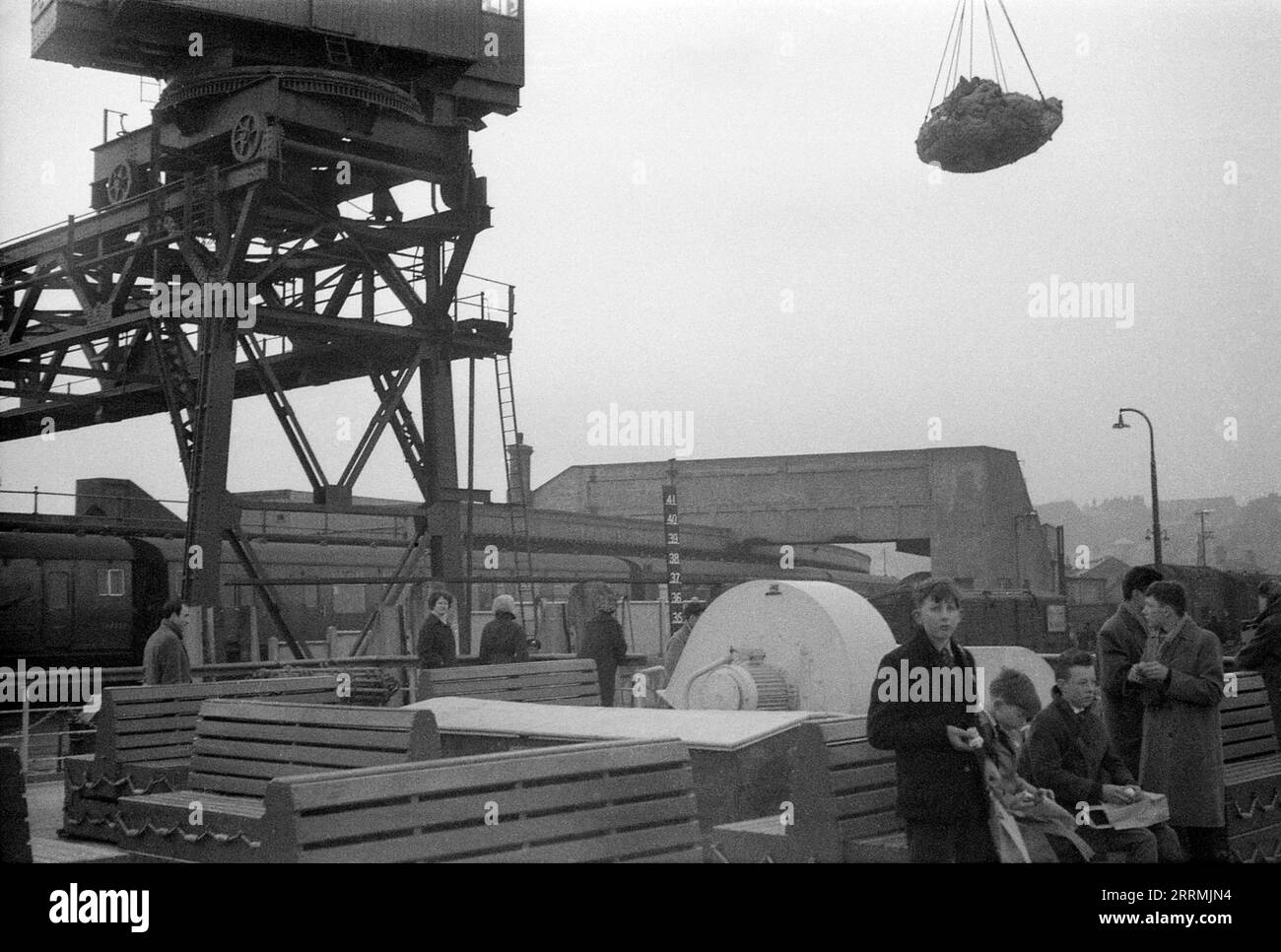 Kent. c.1960 – Passengers on the deck of a French cross-channel ferry at Folkestone Harbour, next to the Folkestone Harbour Railway Station on the harbour arm. A train is at the station. Cargo is being transferred onto the ferry by a quay crane. A group of teenage boys are sitting on a bench, eating lunch. The benches double as life rafts. Stock Photo