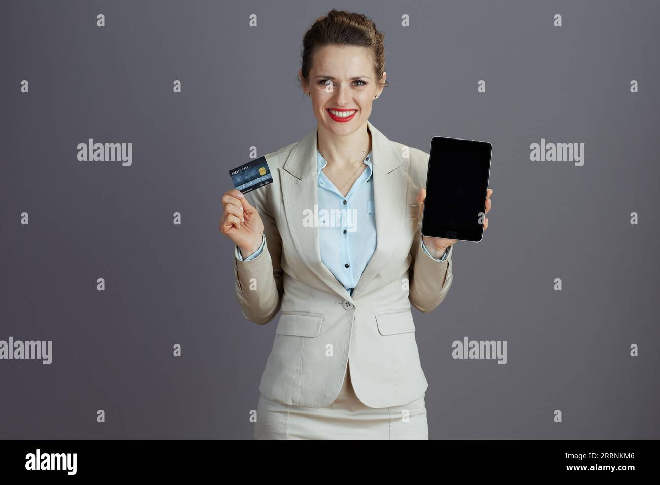 smiling modern female employee in a light business suit with digital tablet and credit card isolated on grey background. Stock Photo
