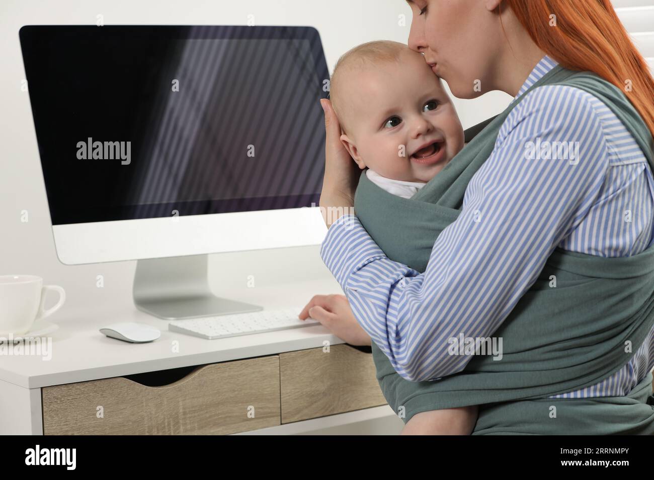Mother holding her child in sling (baby carrier) while using computer ...