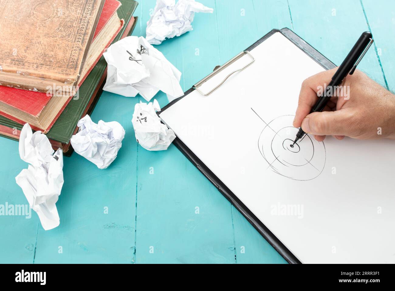Male hand drawing a target on a white clipboard with a black pen next to vintage books and