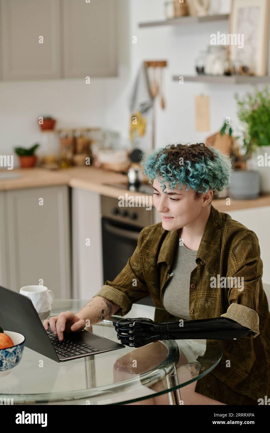 Vertical image of woman with prosthetic arm paying for her purchase online using laptop while sitting in the kitchen Stock Photo