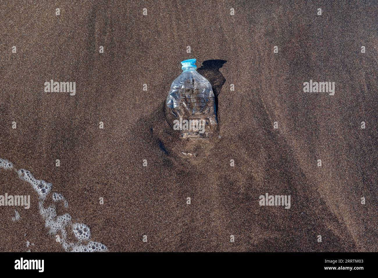 Small squashed plastic water bottle washed on the pristine shores with black volcanic sand of the island of Tenerife, marine debris contaminating Stock Photo