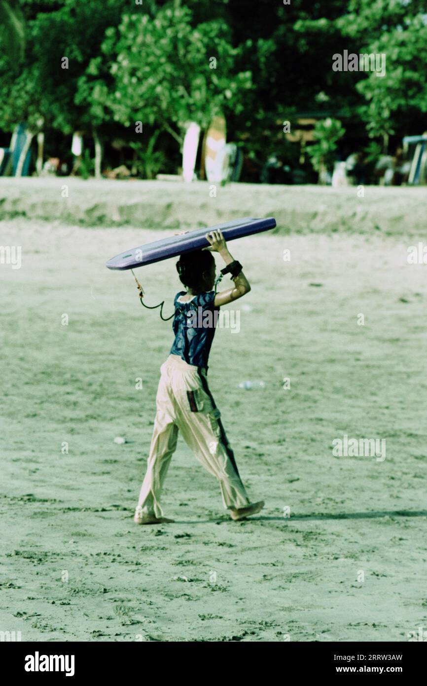 Young boy carrying a surfboard on the beach on the island of Bali Stock Photo