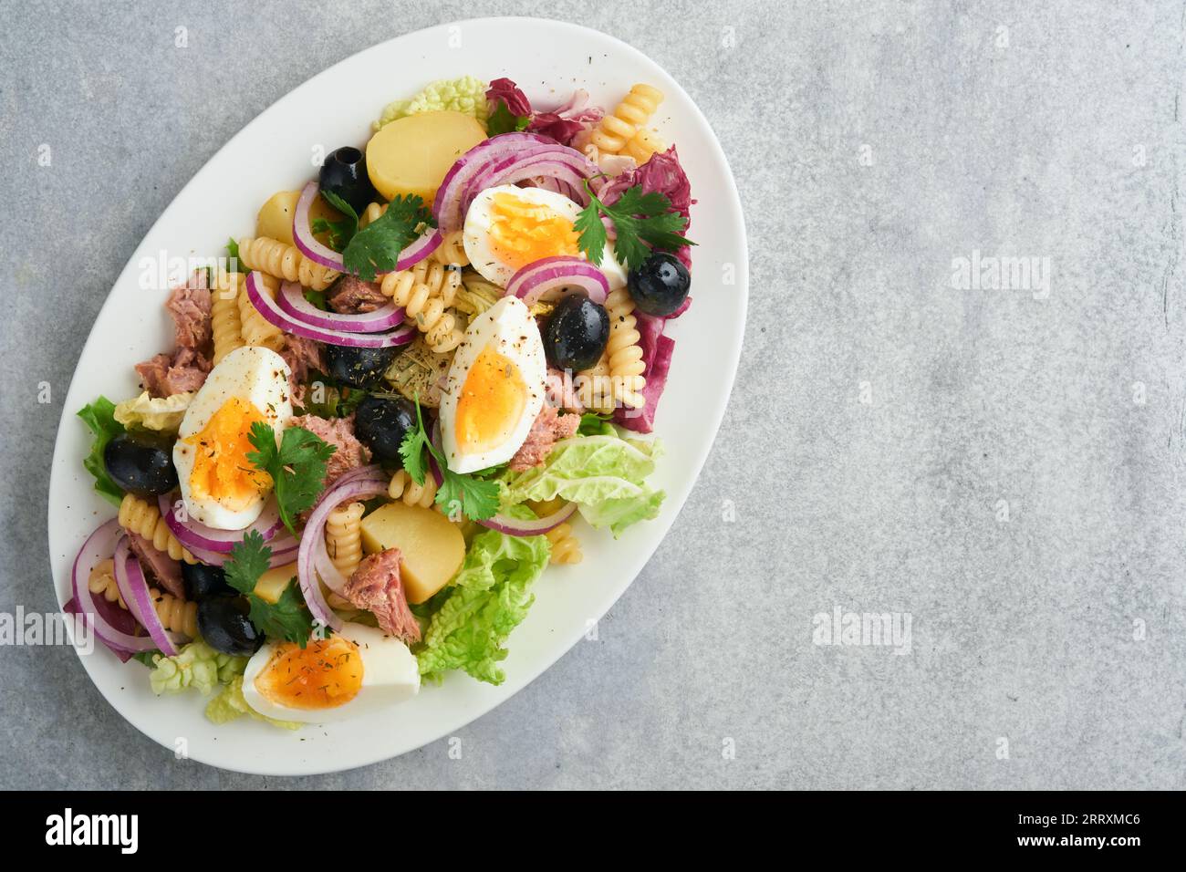 Tuna salad with pasta, eggs, potatoes, olives, red onions and sauce in white plate on old light gray concrete table background. Nicoise salad. French Stock Photo