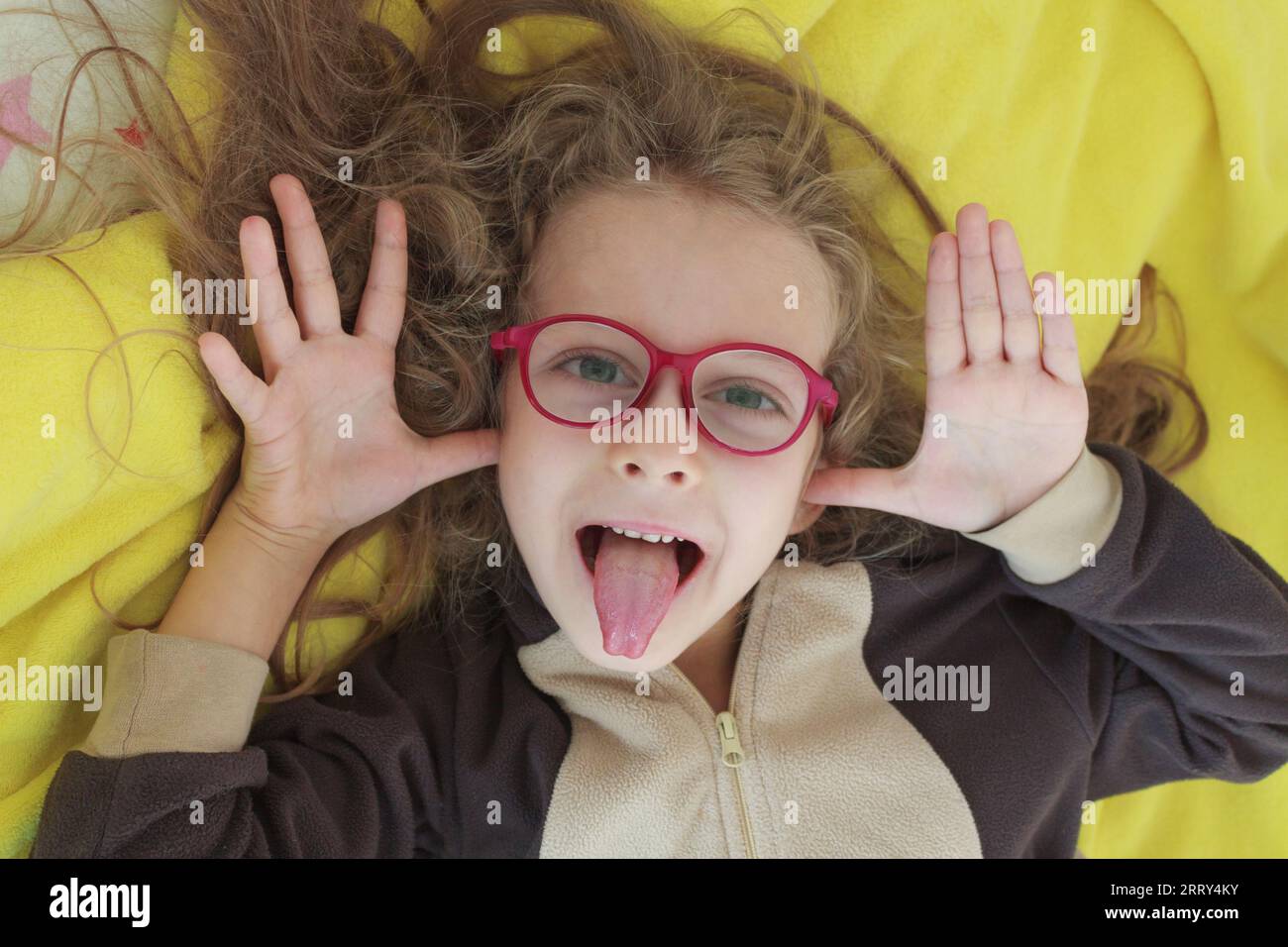 A cheerful girl in glasses makes faces, teases and shows her tongue with her hands in the form of ears, close-up. Stock Photo