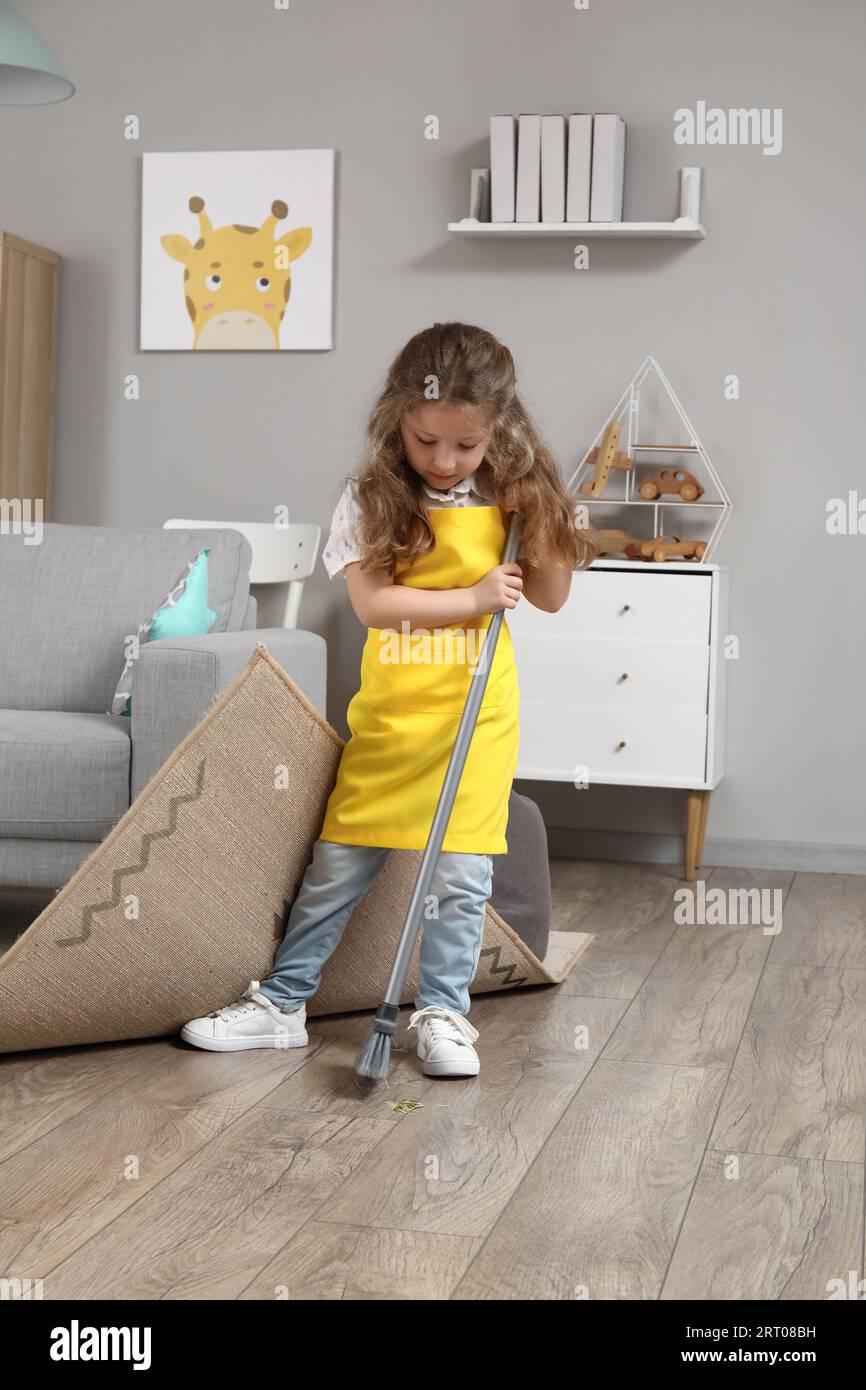 Cute little girl sweeping floor under carpet with broom at home Stock