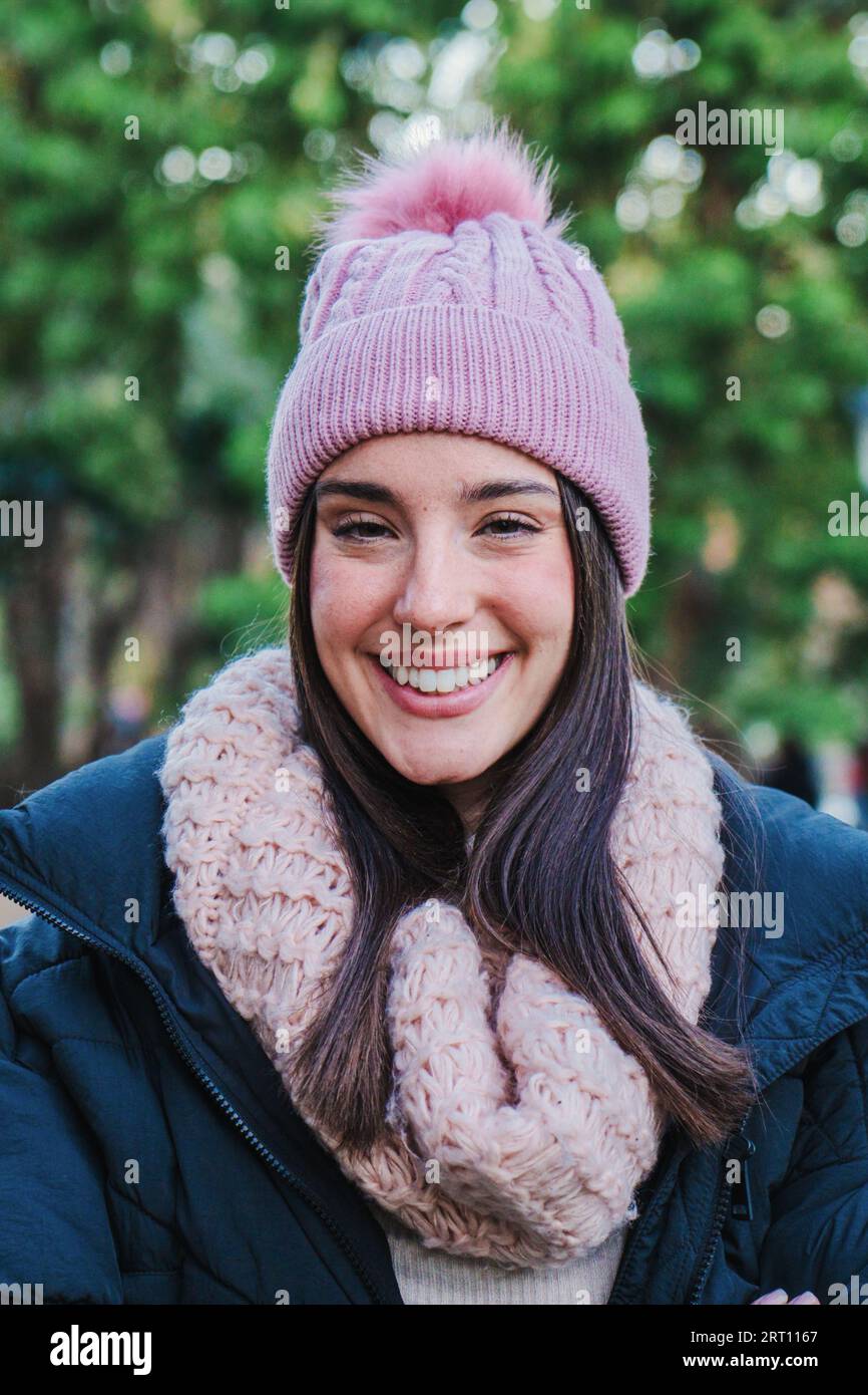 Vertical individual close up portrait of caucasian young woman smiling and looking at camera. Front view of happy teenage brunette girl with autumn clothes, hat, coat and scarf standing at park. High quality photo Stock Photo