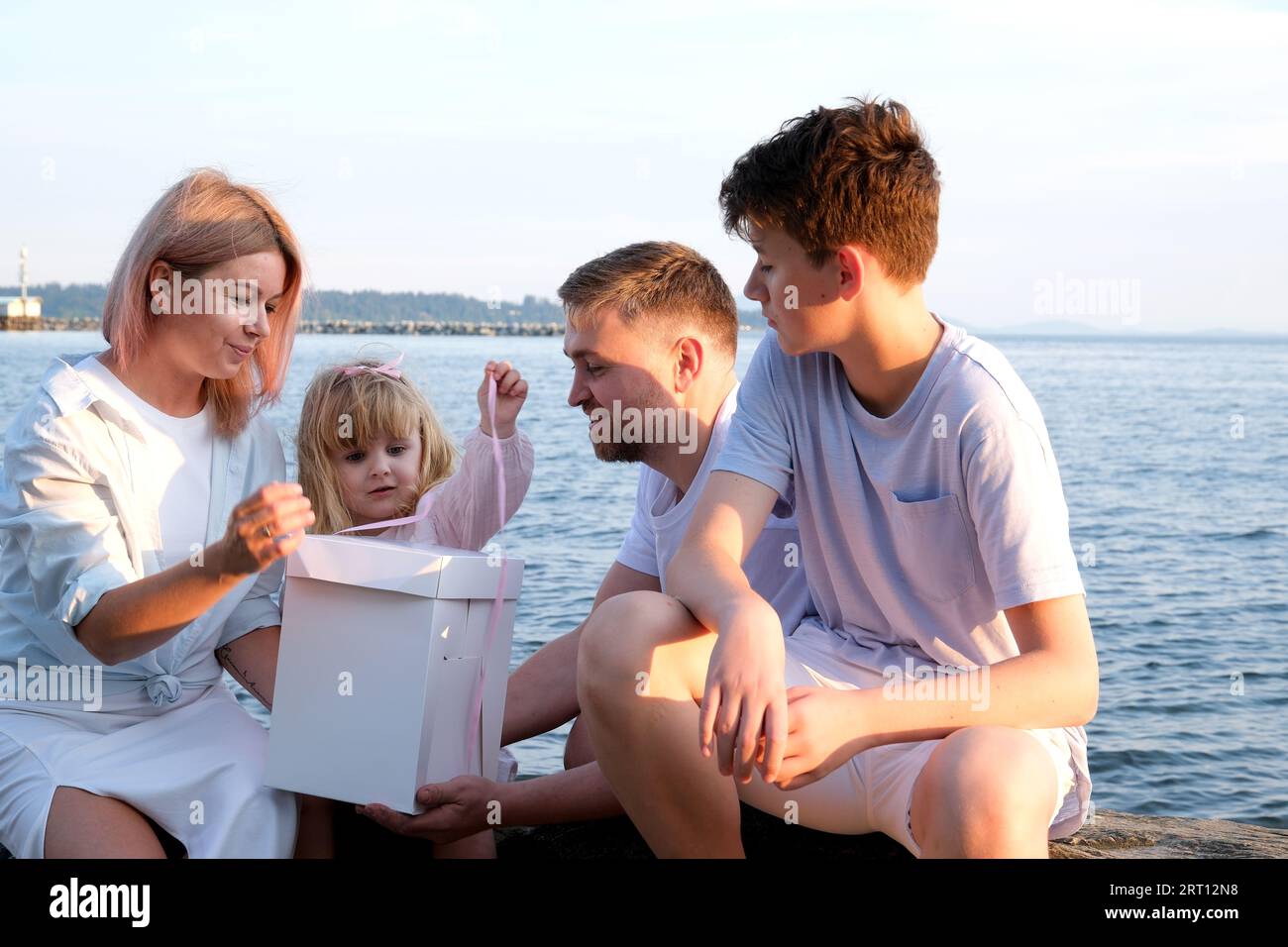 Happy family celebrating little girl's birthday on the seashore mom brings a box with cake girl opens a gift dad mom brother and sister on the ocean shore. on a sunny day Stock Photo