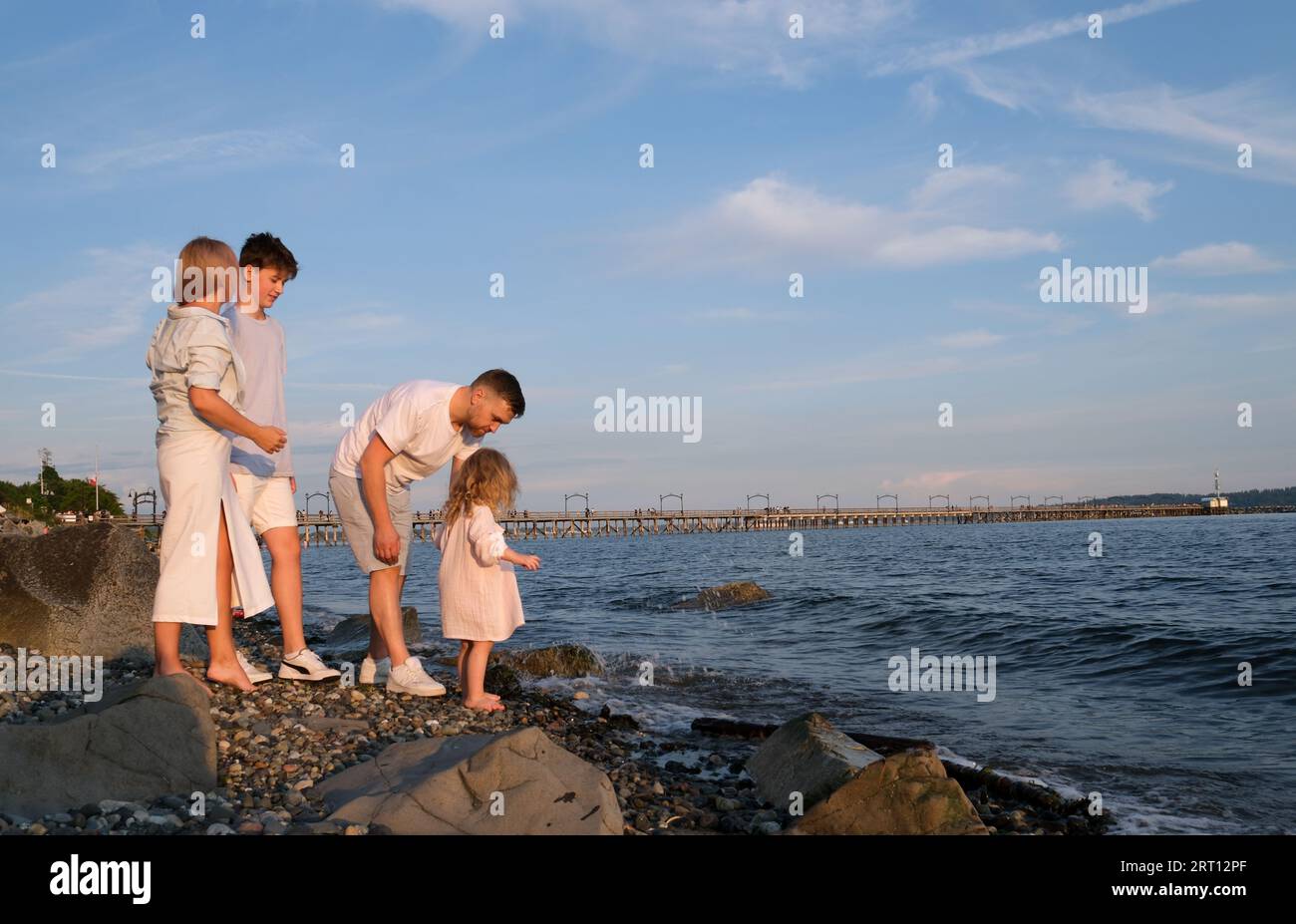 Happy family celebrating little girl's birthday on the seashore mom brings a box with cake girl opens a gift dad mom brother and sister on the ocean shore. on a sunny day Stock Photo