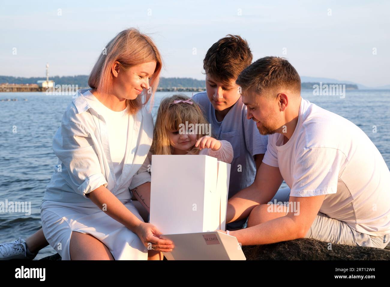 Happy family celebrating little girl's birthday on the seashore mom brings a box with cake girl opens a gift dad mom brother and sister on the ocean shore. on a sunny day Stock Photo