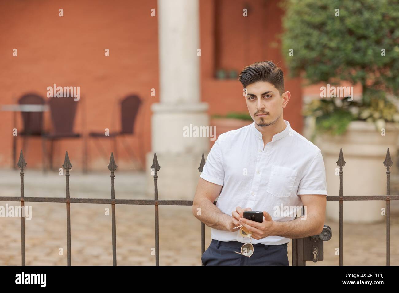 Young man with white short-sleeved shirt holding smartphone and glasses with both hands looking at camera in urban scene with mediterranean flair Stock Photo