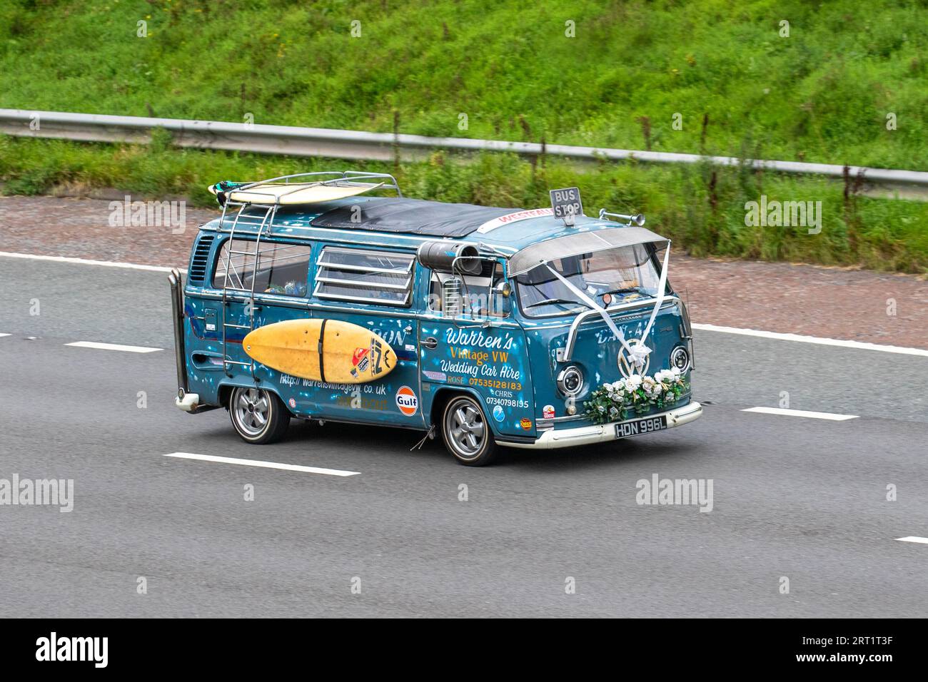 Warren's Vintage VW. 1973 70s seventies Blue Custom Volkswagen Motor Caravan LCV Petrol 1584 cc. Bay Window Westfalia Camper Van a curious wedding hire car with hippie decorations, white ribbons & surfboard; travelling at speed on the M6 motorway in Greater Manchester, UK Stock Photo