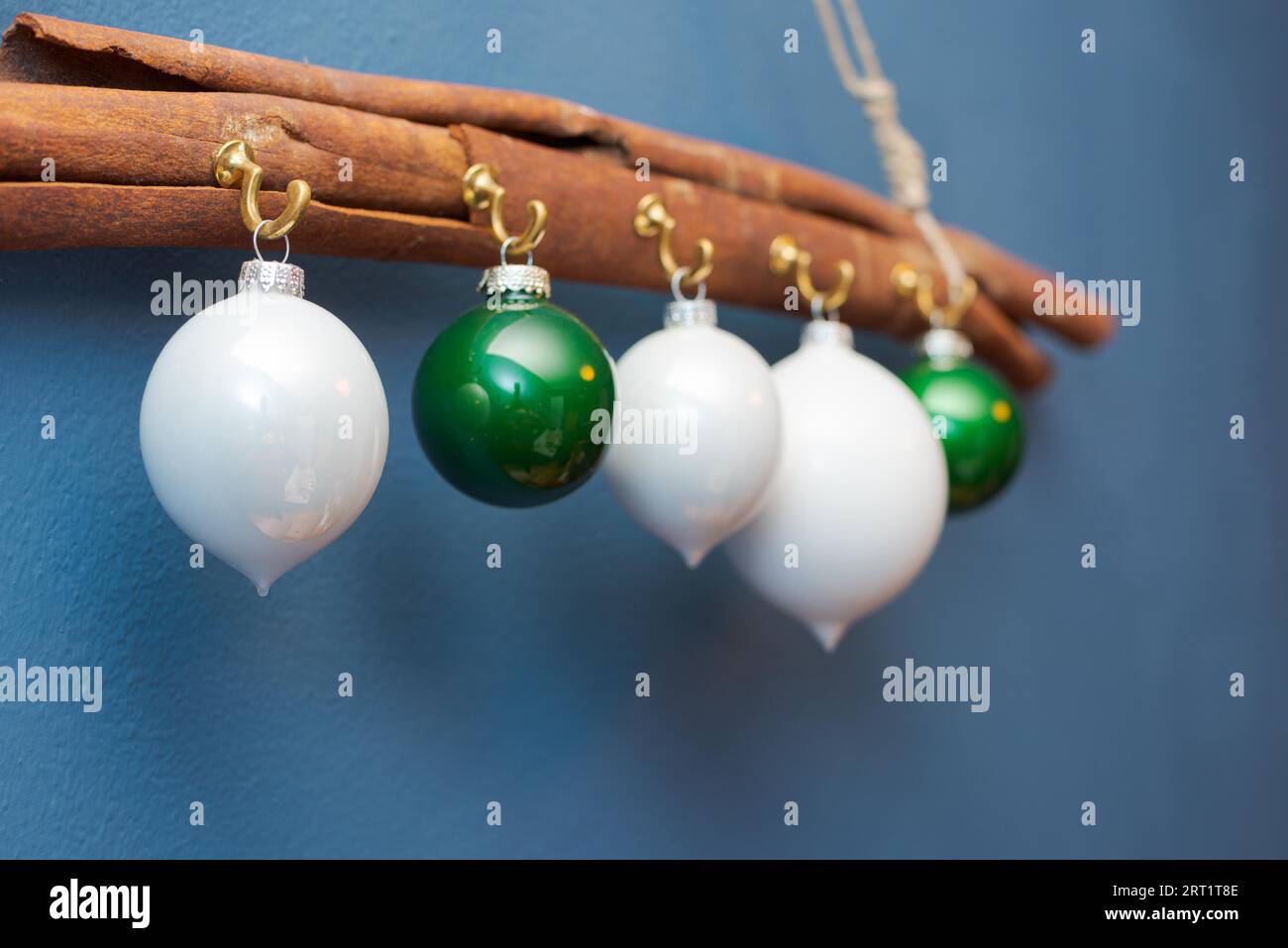 White and green christmas ornament balls hanging on hooks on big cinnamon stick on cord against blue painted wall Stock Photo