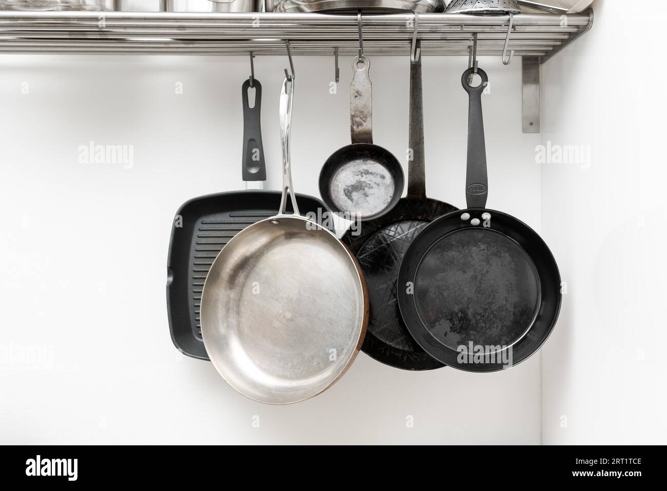 Various pans in different sizes and forms for cooking and frying hanging on metal hooks from shelf in kitchen with white wall in background Stock Photo Alamy