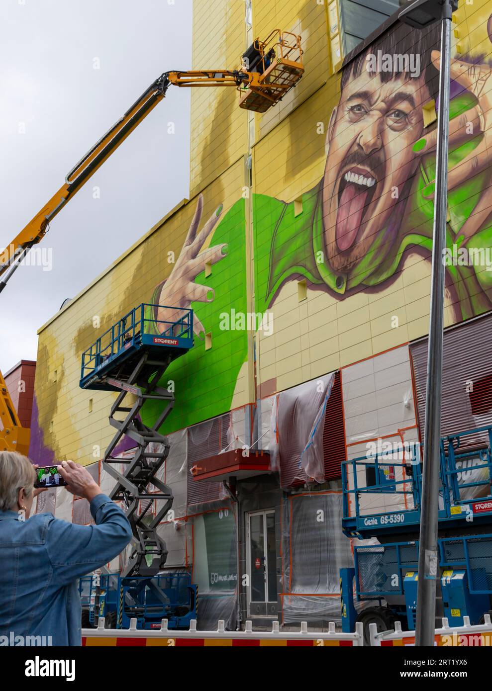 A mural depicting Käärijä being painted, from aerial work platforms, on a Prisma hypermarket wall in Tikkurila. A bypasser is taking a picture. Stock Photo