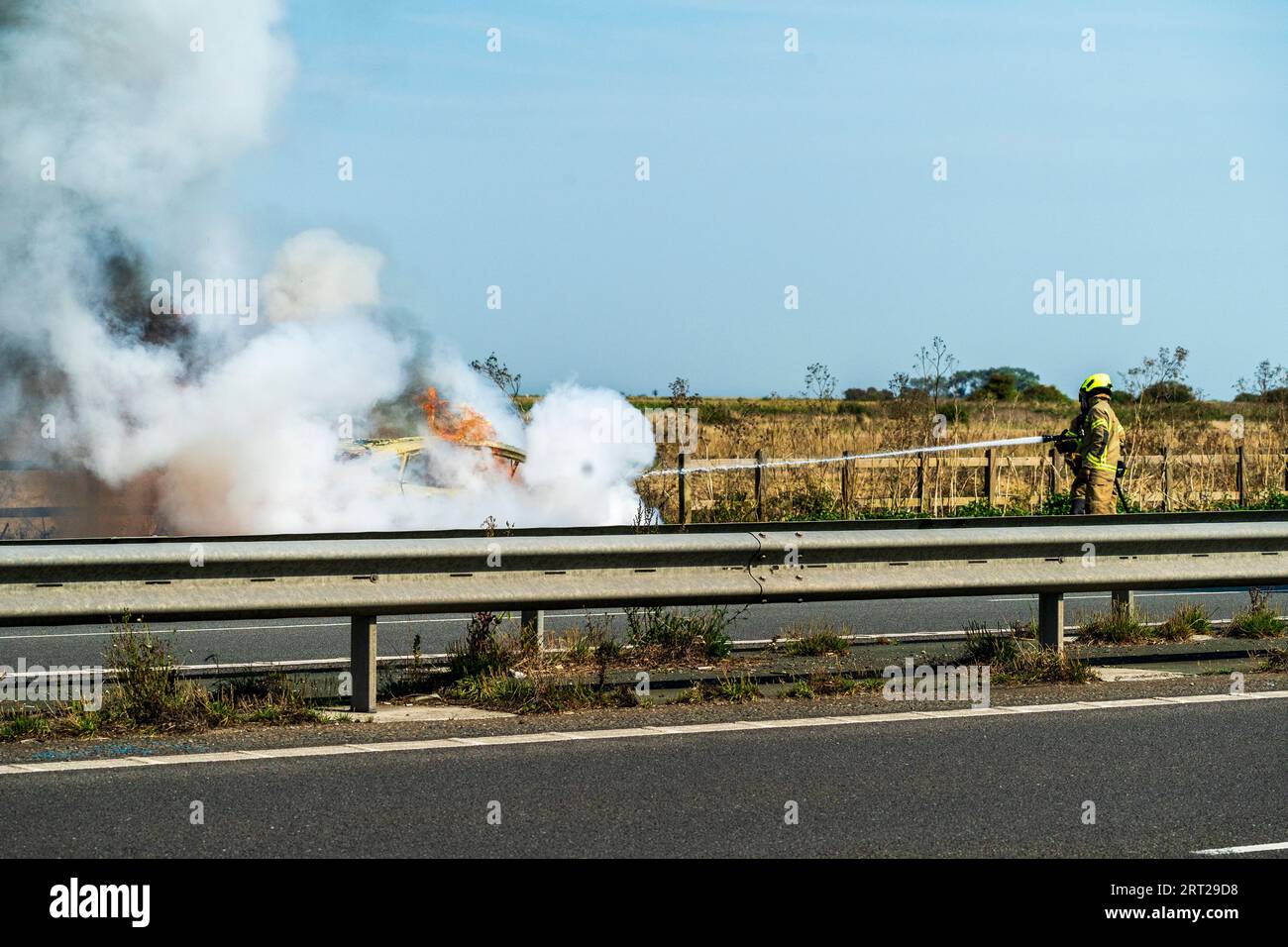 Fireman attending a car fire on a layby on a major road, A299 in the kent countryside. Fireman spraying the fire with water and chemical mix from a hose pipe causing lots of white smoke to billow from the white car. Stock Photo