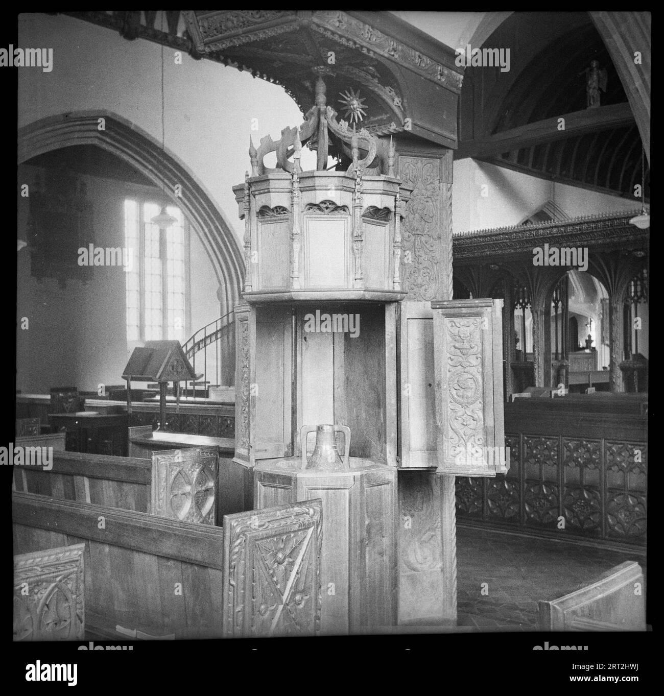 St James' Church, Swimbridge, North Devon, Devon, 1940-1962. Detail of a lead-lined Renaissance font with wooden casing that resembles a pulpit. The font has three tiers, each of which are panelled. The lower tier has plain panels, while the middle tier has decorated panels, of which some hinge open to reveal to top of the font, as seen in the image. The top panels are also plain, but are topped with pinnacles. Above this is an open crown over and a large rectangular canopy. In the background is a chancel screen which crosses the church and was restored in 1880, and above this is an angel stat Stock Photo