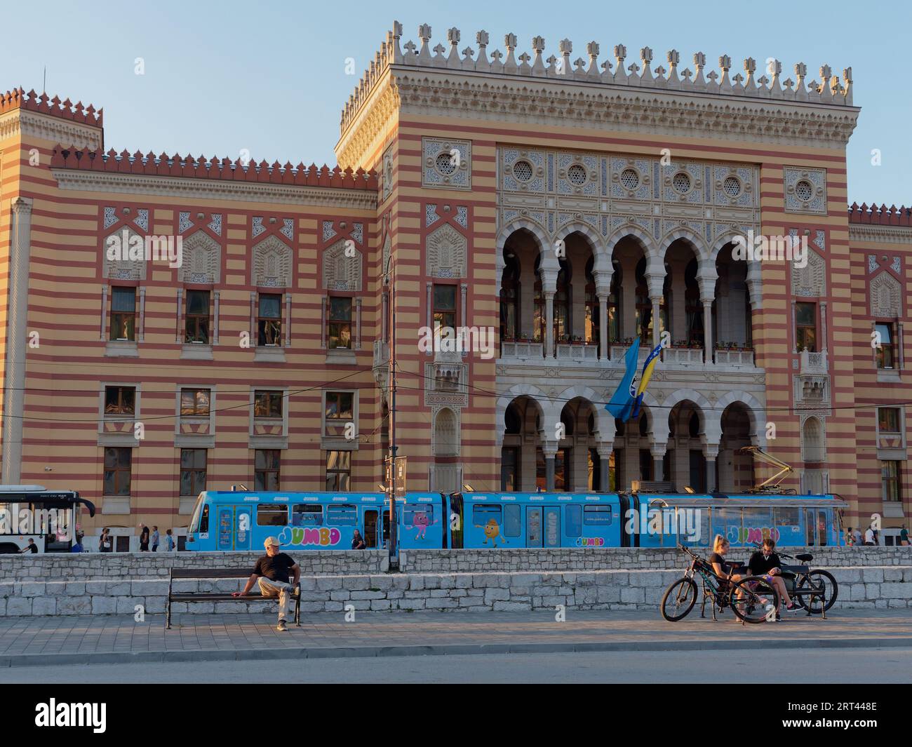Man sits on a bench with City Hall and blue  Tram behind on a summers evening in the city of Sarajevo, Bosnia and Herzegovina, September 10, 2023 Stock Photo