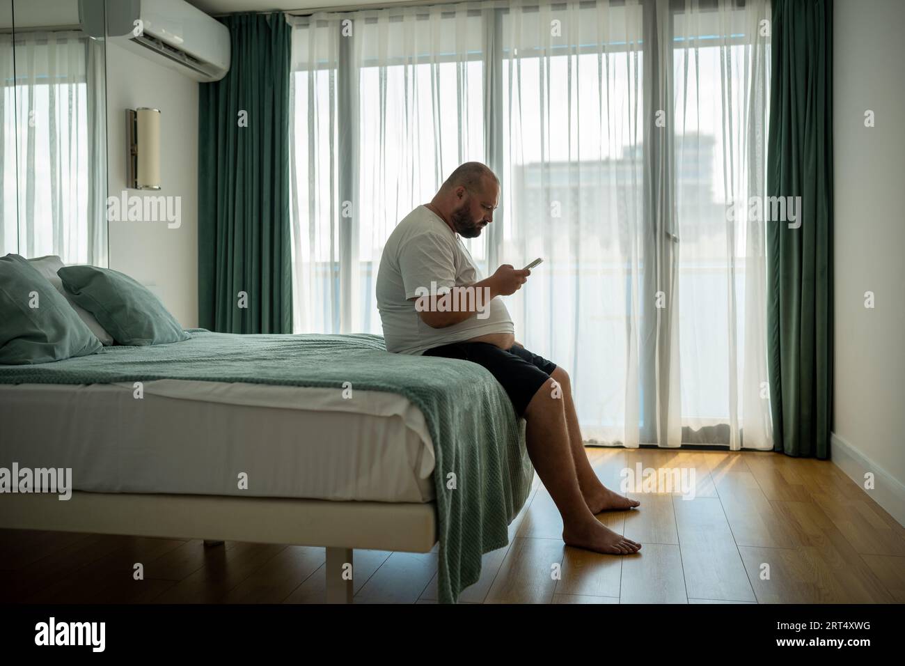 Overweight guy hanging in smartphone on resort sitting in bed in hotel room on vacation. Stock Photo