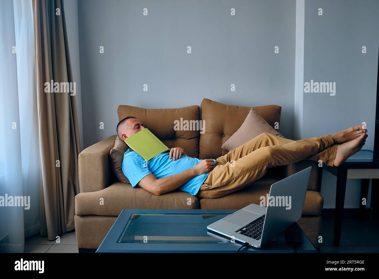 Young adult caucasian man felt asleep on a couch dropped book on face. Tired man sleeps on sofa Stock Photo